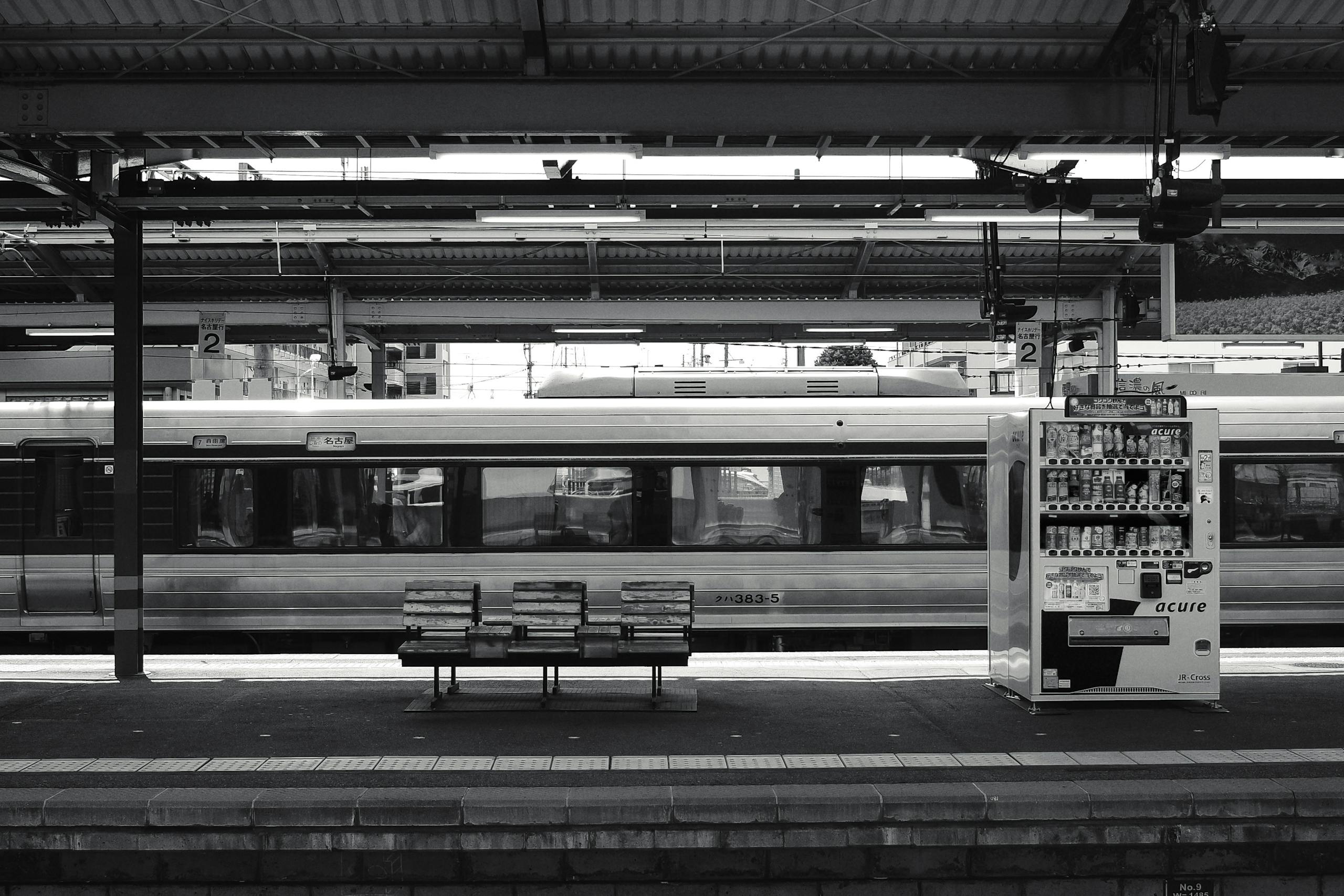 Black and white photo of a Nagano train station with vending machine and benches.